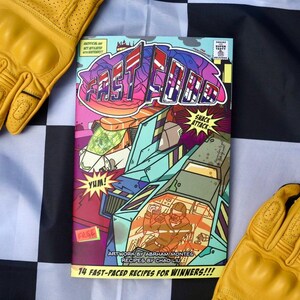 May include: A comic book cover titled "Fast Food" with a colorful, futuristic design. The cover features illustrations of vehicles and a pilot, with text including "Snack Attack" and "14 Fast-Paced Recipes for Winners!!!". Yellow gloves are on either side.
