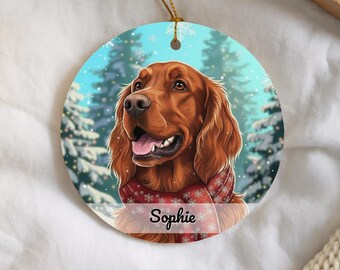 Op de afbeelding: Een rond keramieken ornament met een portret van een bruine Ierse Setter die een rode sjaal met witte sneeuwvlokpatronen draagt. De achtergrond toont een besneeuwd boslandschap met dennenbomen. De naam Sophie is in het zwart afgedrukt.