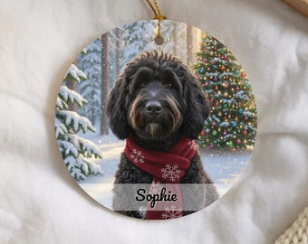 May include: A round ceramic Christmas ornament featuring a black curly-haired dog wearing a red scarf with white snowflake patterns. The background shows a snowy forest and a decorated Christmas tree. The name Sophie is printed in black script.