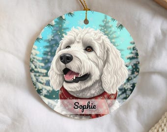 May include: A round ceramic ornament featuring a white curly-haired dog wearing a red scarf with a snowflake pattern. The background shows a snowy forest scene with pine trees. The name Sophie is printed in black script across the lower section.