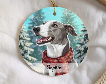 Può includere: Un ornamento rotondo in ceramica con un'illustrazione digitale di un cane grigio e bianco che indossa una sciarpa rossa con motivo a fiocchi di neve. Lo sfondo mostra una foresta innevata con fiocchi che cadono. Il nome Sophie è stampato in nero.