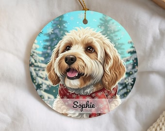 May include: A round Christmas ornament featuring a smiling dog with light brown fur, wearing a red scarf with white snowflake patterns. The background shows a snowy forest scene with snowflakes and the name "Sophie" on the scarf.