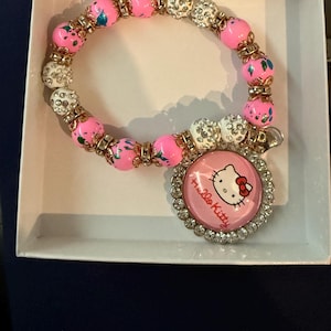 Op de afbeelding: Roze en witte kralen armband met een Hello Kitty bedel. De kralen zijn versierd met gouden accenten en witte strass steentjes.