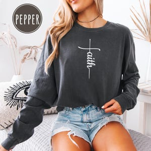 May include: Dark gray long-sleeve sweatshirt with the word "faith" in white script, designed as a cross. The sweatshirt is paired with denim shorts. The image includes a round logo with the word "pepper".