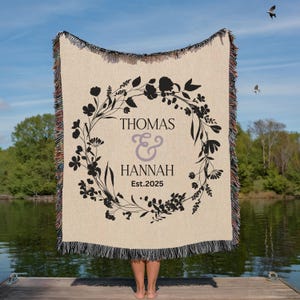 May include: A beige woven throw blanket with a black floral wreath design. The text "THOMAS & HANNAH Est.2025" is centered within the wreath. The blanket has fringed edges and is held up against a blue sky.