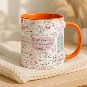May include: A ceramic mug with an orange interior and handle, featuring inspirational quotes and floral designs. The mug has a white exterior with text in various fonts and colours, including pink, green, and brown. The mug is sitting on a cream-coloured knitted surface.