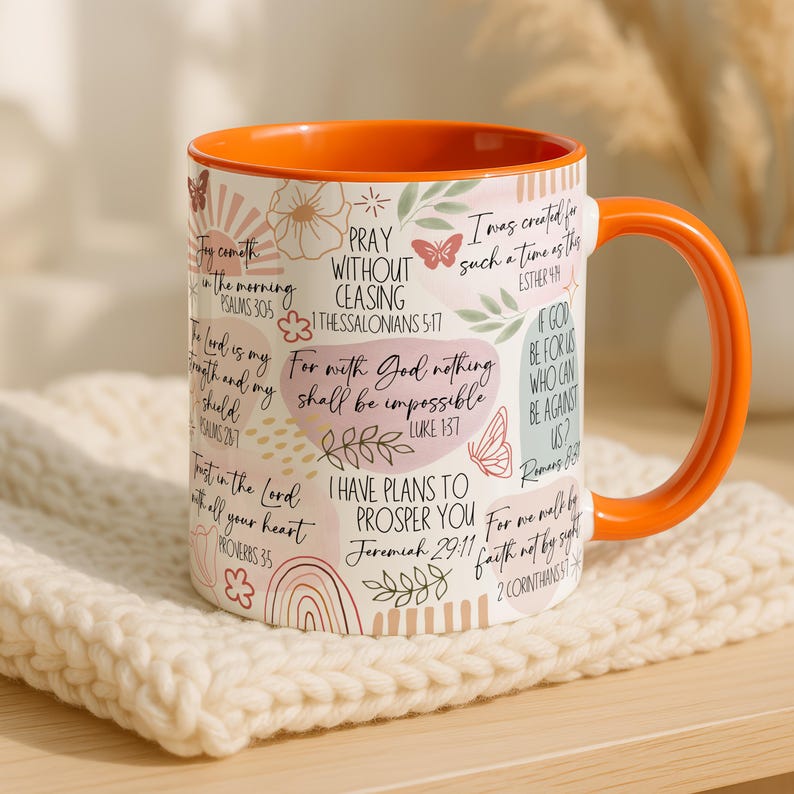 May include: A ceramic mug with an orange interior and handle, featuring inspirational quotes and floral designs. The mug has a white exterior with text in various fonts and colours, including pink, green, and brown. The mug is sitting on a cream-coloured knitted surface.