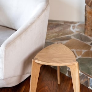 May include: A wooden side table with a triangular top and three legs. The table is made of light-coloured wood and has a minimalist design. The table is next to a cream-coloured upholstered chair.