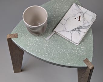 May include: A modern, triangular side table with a speckled, mint-green top and wooden legs. A beige ceramic cup and a marble-patterned notebook with the letter "A" rest on the table. The table's design is minimalist and stylish.