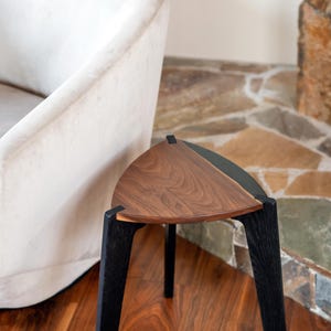 May include: A modern, triangular side table with a walnut wood top and black legs. The table has a unique design and is placed on a hardwood floor. The background includes a white armchair and a stone wall.