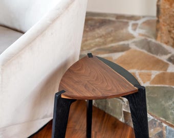 May include: A modern, triangular side table with a walnut wood top and black legs. The table has a unique design and is placed on a hardwood floor. The background includes a white armchair and a stone wall.