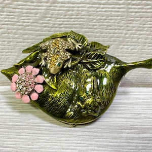 May include: A decorative trinket box shaped like a leaf, with a green and gold finish. The box features a jeweled frog and a pink flower with crystal accents. A smaller frog is also visible.