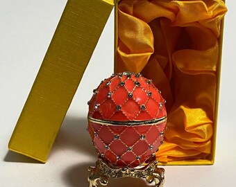 May include: A red and gold jeweled egg trinket box on a gold stand. The egg is decorated with a diamond pattern and is in a yellow box with a satin lining.