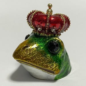 May include: A decorative frog figurine with a red and gold crown. The frog's head is green and gold with black gem eyes. The crown is red with gold trim and small clear gems. The figurine is on a white surface.