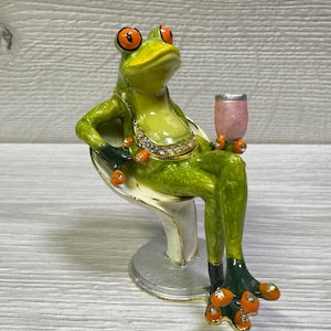 May include: A whimsical green frog figurine with orange eyes and accents, seated on a white and gold chair. The frog holds a pink glass and wears a jeweled collar. The figurine has a silver base.