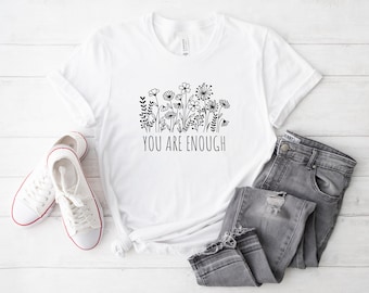May include: A white t-shirt with a black floral design and the words "YOU ARE ENOUGH". The shirt is paired with gray jeans and white sneakers with red trim. The image is set against a white wooden background.