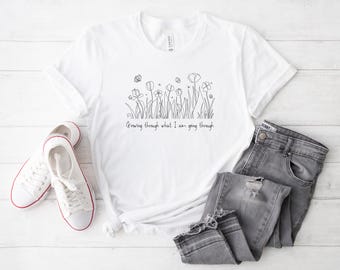May include: White t-shirt with a black line drawing of flowers and the phrase "Growing through what I am going through." The shirt is paired with gray jeans and white sneakers with red trim.
