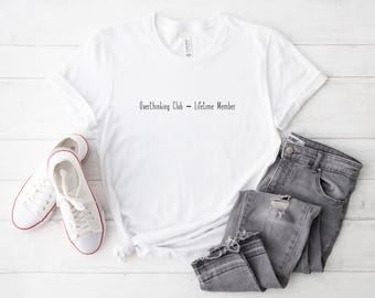 May include: A white t-shirt with the text "Overthinking Club - Lifetime Member" in black. The shirt is paired with gray jeans and white sneakers with red trim, all arranged on a light wood-grain surface.