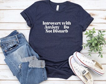 May include: A navy blue t-shirt with white text that reads "Introvert with Anxiety - Do Not Disturb". The shirt is laying on a white wooden surface with a pair of blue jeans and white sneakers.