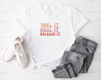 May include: White t-shirt with the text "Feel it. Heal it. Release it." in a colorful, retro font.