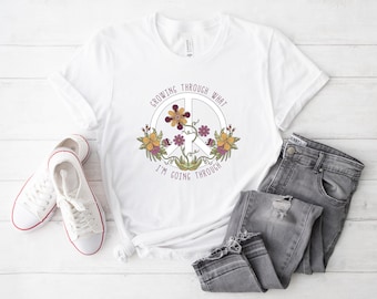 May include: White t-shirt with a peace sign design featuring flowers in shades of yellow, purple, and brown. The text "Growing Through What I'm Going Through" is printed around the peace sign. A pair of white sneakers and gray jeans are next to the shirt.