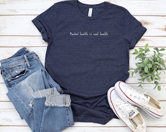 May include: A navy blue t-shirt with the text "Mental health is real health" in white lettering. The t-shirt is laid flat on a white wooden surface with a pair of blue jeans and white sneakers.