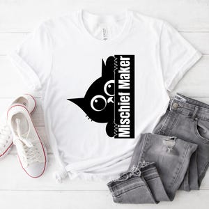Peut inclure: T-shirt blanc avec un chat noir qui regarde derrière une pancarte qui dit "Mischief Maker".