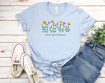 May include: Light blue t-shirt with a floral design and the text "Feel it. Heal it. Release it." The shirt is paired with a pair of white canvas shoes and blue jeans.