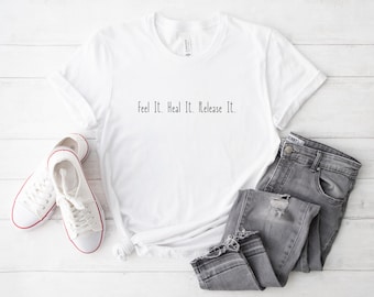 May include: White t-shirt with the text "Feel It. Heal It. Release It." printed on the front. The shirt is laying on a white wooden surface with a pair of white sneakers and a pair of gray jeans.