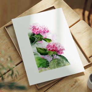 Puede incluir: Una impresión en acuarela de flores de hortensia rosa con hojas verdes. La obra de arte está sobre papel blanco, sobre una superficie de madera. Una taza de café está en la esquina inferior derecha. La impresión está en un marco de madera.
