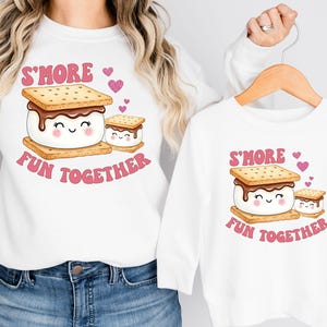 May include: White sweatshirts with a matching graphic of a s'more with the text "S'MORE FUN TOGETHER" in pink. The s'more design includes a smiling marshmallow, chocolate, and graham crackers. One sweatshirt is adult size, the other is child size.