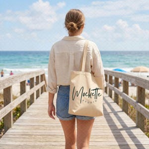 Puede incluir: Un bolso de tela beige con el texto negro Mockette Studio impreso en la parte delantera. El bolso tiene dos correas para los hombros y se lleva al costado. El diseño es sencillo y funcional, hecho de un material de tela de color claro.