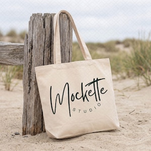 May include: A beige canvas tote bag with two shoulder straps, featuring the dark green text Mockette STUDIO printed on the front. The bag is constructed from a light-colored fabric and displays a simple, rectangular design with a flat base.