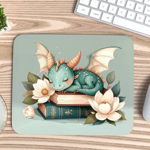 Puede incluir: Una alfombrilla de ratón con un dragón turquesa durmiendo sobre libros y flores blancas. El dragón tiene cuernos y alas naranjas. La alfombrilla está sobre un escritorio de madera, con teclado y ratón.