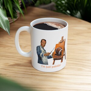 May include: White ceramic coffee mug with a graphic of a man painting a football on a canvas. The text "I'm just paintin'" is printed on the bottom of the mug.