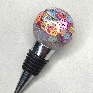 May include: A decorative bottle stopper with a clear, round top filled with colorful flower-shaped confetti. The stopper has a silver metal base and a black rubber seal.