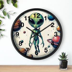 May include: A round wall clock with a black frame featuring a green alien illustration. The clock face has a cream background with planets and stars. Black hands indicate the time. The alien is in the center.