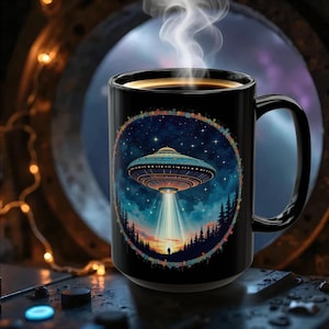 May include: Black ceramic coffee mug featuring a colorful illustration of a UFO hovering over a forest at night, with a starry sky background. The mug has a large handle and is filled with hot coffee, with steam rising from the top.