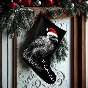 May include: Black Christmas stocking with a grayscale raven wearing a Santa hat. The raven is perched on a branch, with the text "F-CAW-F" below. Festive holiday decor includes red baubles and evergreen branches.
