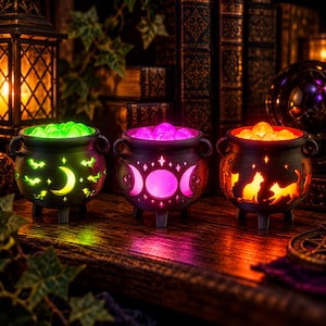 May include: Three black ceramic cauldrons with glowing interiors. The left cauldron features cutouts of bats and a crescent moon with green light. The center cauldron shows a triple moon symbol with purple light. The right cauldron displays cat silhouettes with orange light.