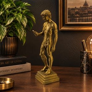 Könnte beinhalten: Eine goldfarbene Statue einer stehenden Figur mit lockigem Haar im klassischen Stil, die ein kleines rechteckiges Gerät in einer Hand hält. Die Figur steht auf einem quadratischen Sockel und hat ein kleines Blattbündel im Lendenbereich.