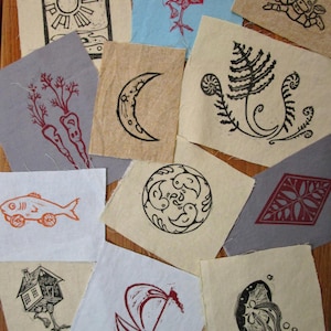 May include: A collection of fabric squares with various linocut designs. Images include a sun, house, moon, fern, fish, birds, and a sailboat. The prints are in black, red, and orange ink on neutral and colored fabric.