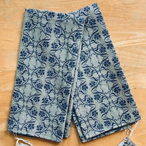 May include: Two folded, light green kitchen towels with a repeating dark blue floral pattern. The towels are made of a textured fabric and have small tags attached. The floral design features stylized flowers and vines.