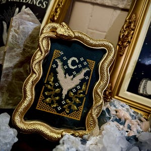 Puede incluir: Un marco dorado en forma de serpiente con un diseño bordado en azul oscuro y dorado de un murciélago y una luna creciente. El marco está sobre un lecho de cristales blancos.