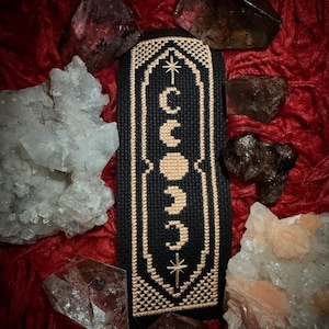 May include: A black and cream cross-stitch bookmark with a design of a crescent moon and stars. The bookmark is surrounded by crystals.