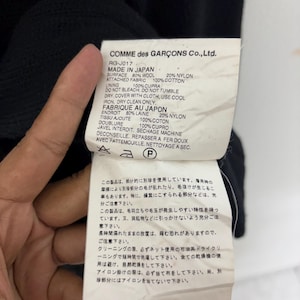 May include: Close-up of a tag from a Comme des Gar&ccedil;ons garment. The tag is white with black text, listing fabric composition: 80% wool, 20% nylon, and 100% cotton. It also includes care instructions and the phrase "Made in Japan."