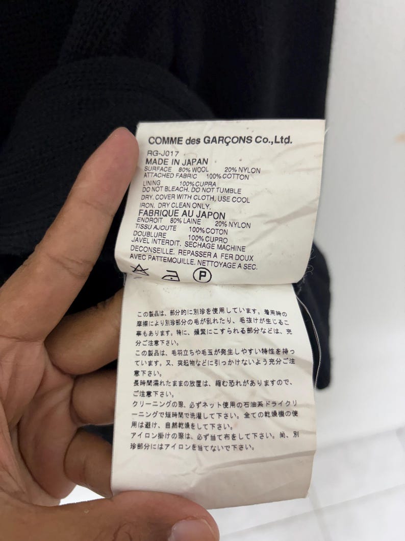May include: Close-up of a tag from a Comme des Gar&ccedil;ons garment. The tag is white with black text, listing fabric composition: 80% wool, 20% nylon, and 100% cotton. It also includes care instructions and the phrase "Made in Japan."