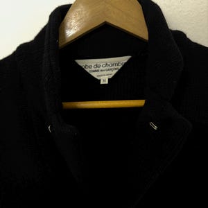 May include: A black jacket with a notched collar, hanging on a wooden hanger. The label reads "robe de chambre COMME des GAR&Ccedil;ONS MADE IN JAPAN M". The jacket has two metal closures and a third at the bottom.