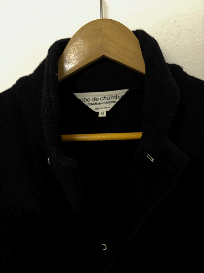 May include: A black jacket with a notched collar, hanging on a wooden hanger. The label reads "robe de chambre COMME des GAR&Ccedil;ONS MADE IN JAPAN M". The jacket has two metal closures and a third at the bottom.