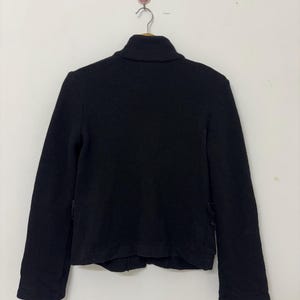 May include: A black jacket with a high collar, hanging on a hook. The jacket has long sleeves and a simple, classic design. The fabric appears to be a medium-weight material, suitable for cooler weather.